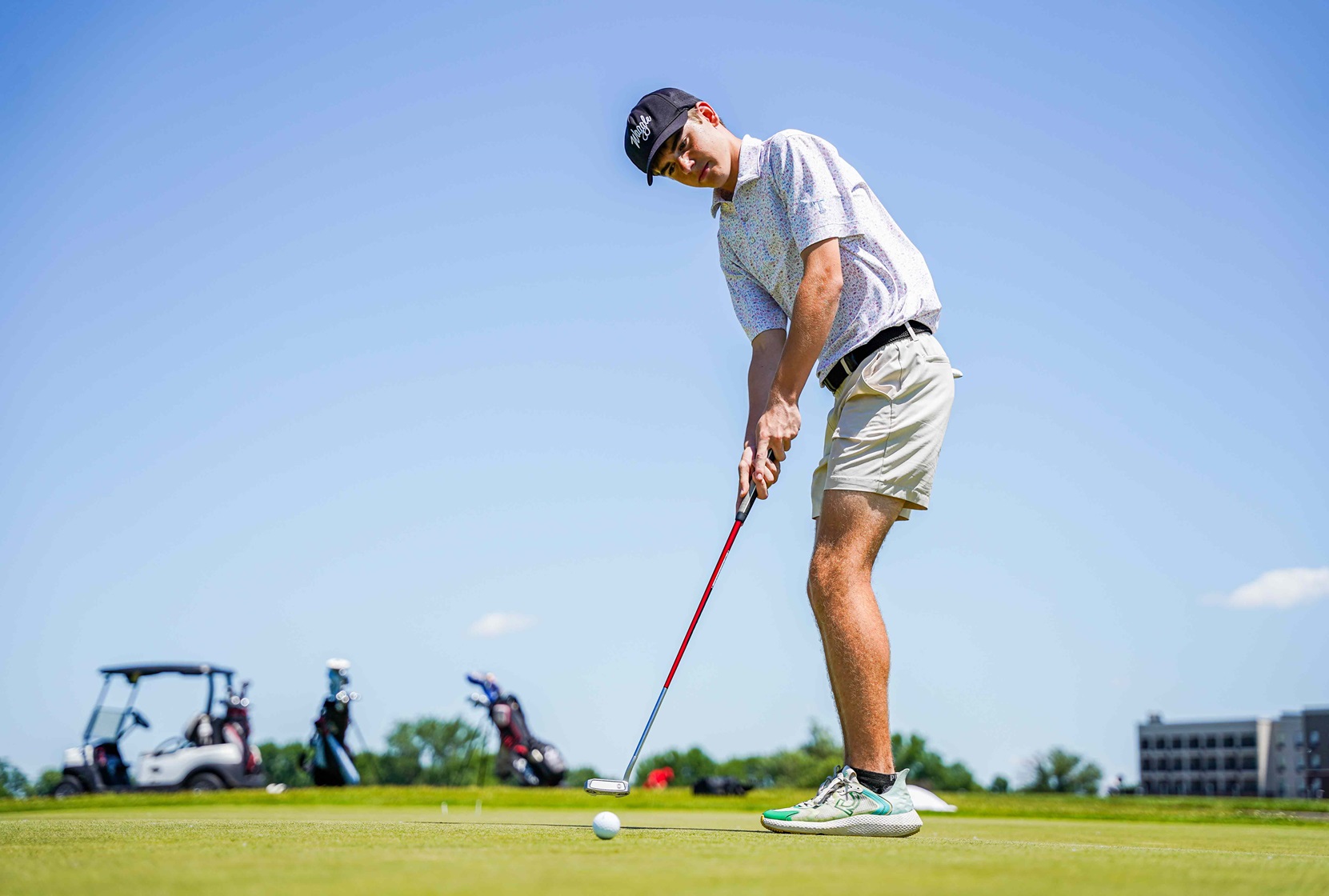 adult golf player outdoors hitting a golf ball in front of a blue sky and golf cart in the distance